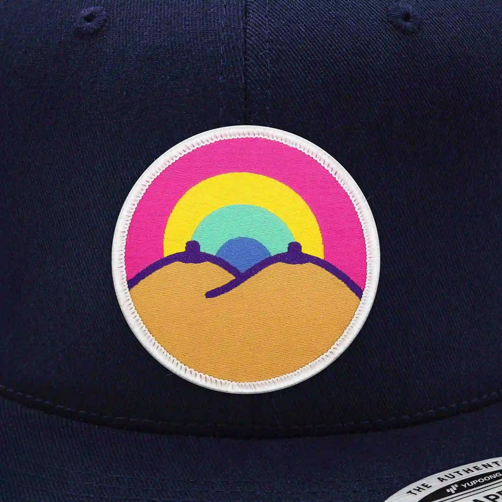 Casquette Flexfit bleu royal snapback avec écusson rond velcro représentant deux collines brunes et un arc-en-ciel multicolore, modèle streetwear personnalisable avec écussons interchangeables.