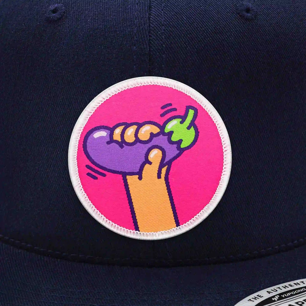 Casquette Flexfit bleu royal snapback avec écusson rond velcro représentant une aubergine violette stylisée sur fond rose, modèle streetwear personnalisable avec écussons interchangeables.