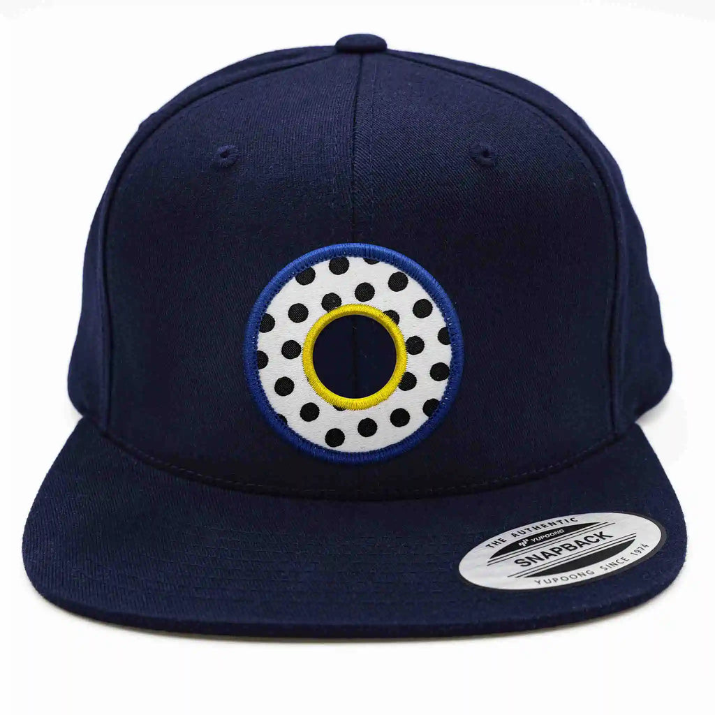 Casquette Flexfit bleue marine snapback avec écusson rond velcro au design Memphis blanc à pois noirs et contour jaune et bleu, modèle personnalisable avec écussons interchangeables.
