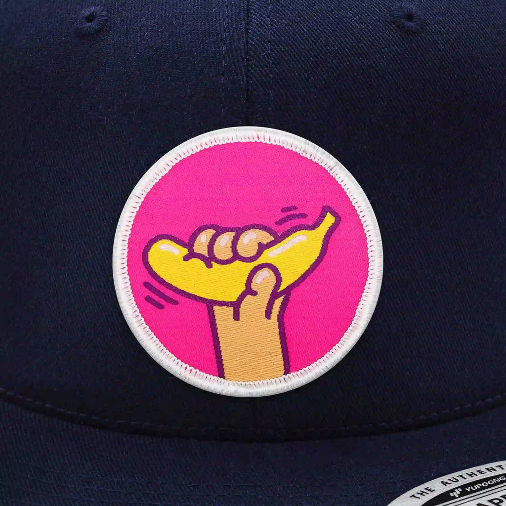 Casquette Flexfit navy snapback avec écusson rond velcro représentant une main tenant une banane jaune sur fond rose, modèle original personnalisable avec écussons interchangeables.