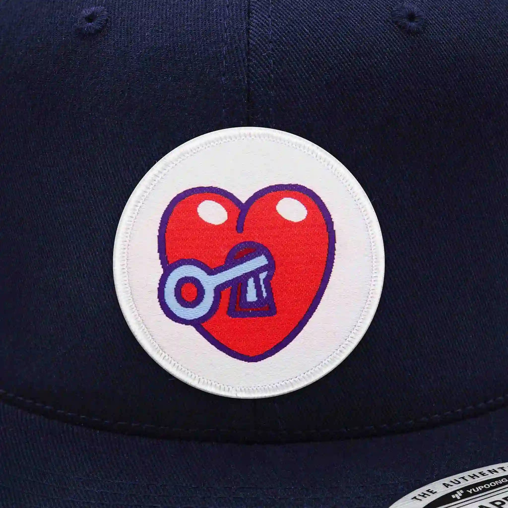 Casquette Flexfit bleu navy snapback avec écusson rond velcro représentant un cœur rouge avec serrure et clé, modèle streetwear personnalisable avec écussons interchangeables.
