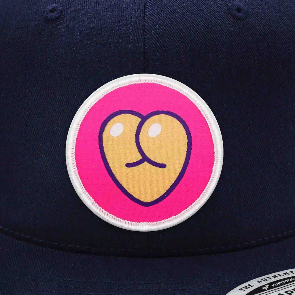 Casquette Flexfit bleu navy snapback avec écusson rond velcro représentant un cœur en forme de fesses stylisées sur fond rose, modèle original et personnalisable avec écussons interchangeables.