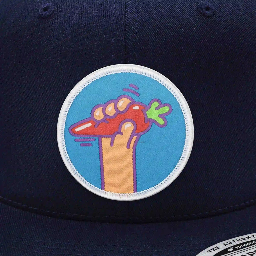 Casquette Flexfit bleu navy snapback avec écusson rond velcro représentant une main tenant une carotte orange sur fond bleu, modèle humoristique personnalisable avec écussons interchangeables.