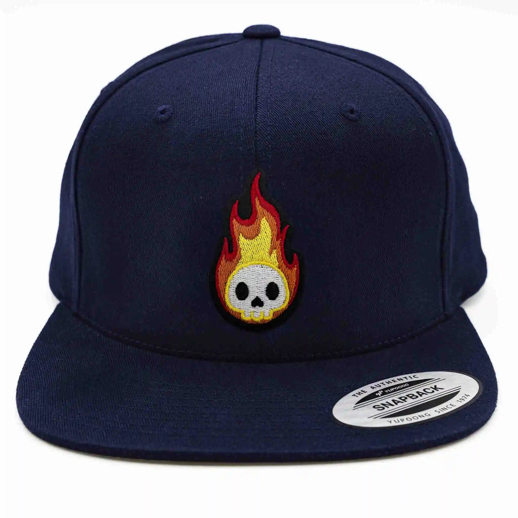 Casquette Flexfit bleue marine snapback avec écusson brodé représentant un crâne blanc entouré de flammes rouges et jaunes, modèle streetwear personnalisable avec velcro et écussons interchangeables.