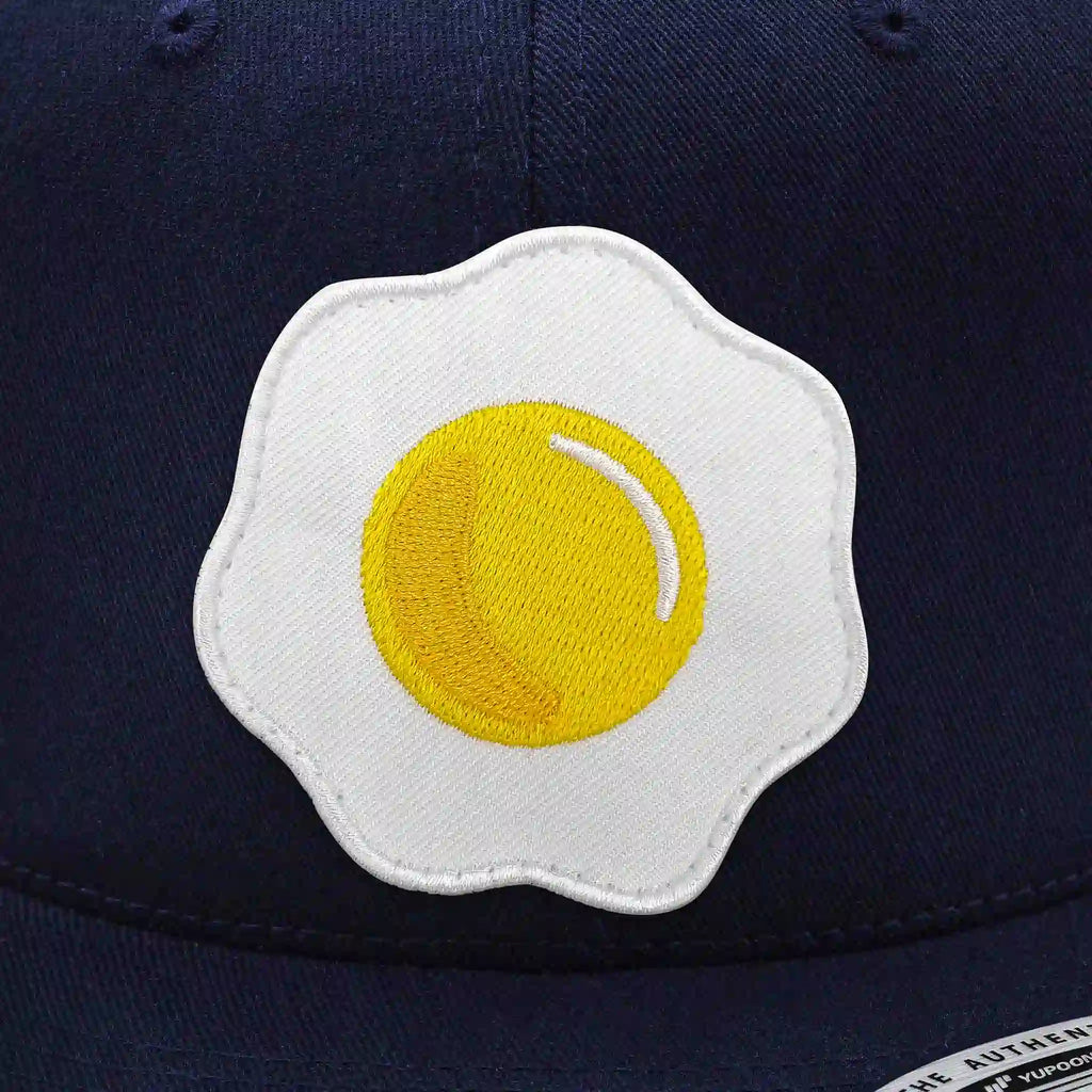 Casquette Flexfit bleu navy snapback avec écusson rond velcro brodé en forme d’œuf au plat jaune et blanc, modèle humoristique personnalisable avec écussons interchangeables.