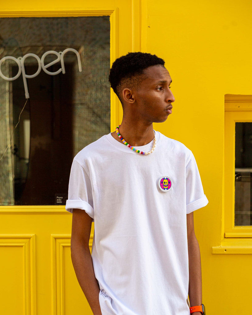 Look complet avec t-shirt blanc et écusson capsule brodé, porté par un homme devant une façade jaune