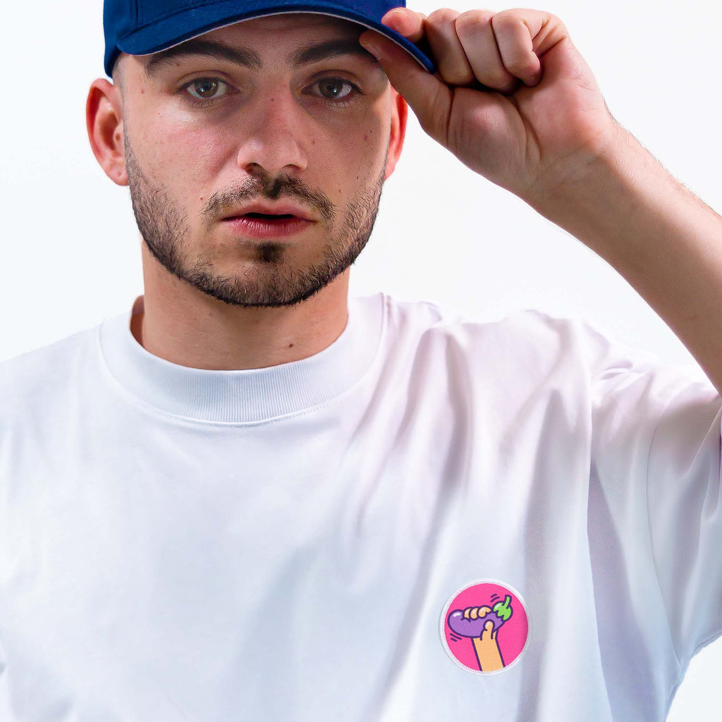 Homme en casquette bleue portant un t-shirt blanc décoré d’un écusson aubergine coloré, style patch thermocollant ou scratch.