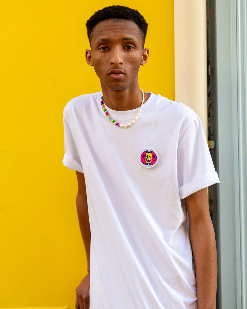 Patch capsule coloré porté sur poitrine d’un t-shirt blanc, modèle accoudé à une façade jaune – idée style street casual