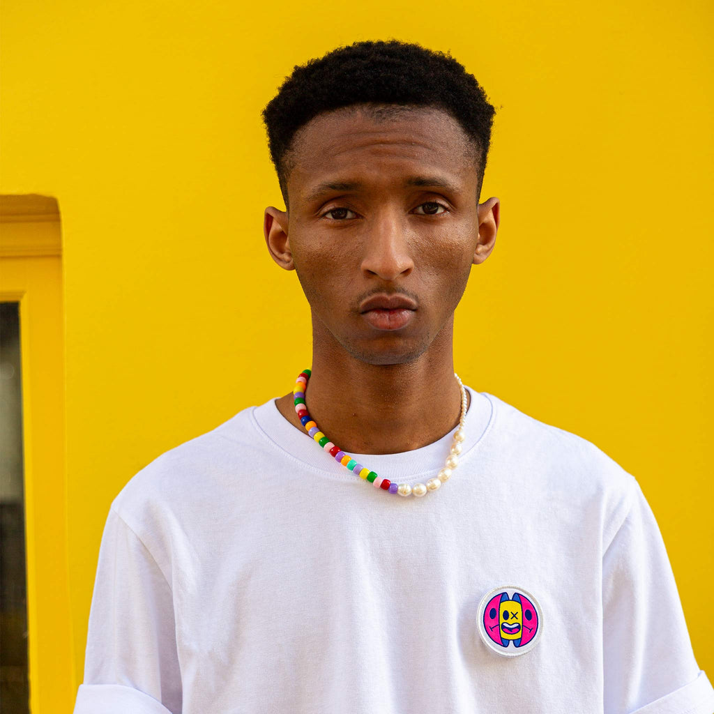 Portrait d’un jeune homme portant un patch capsule brodé sur t-shirt blanc, devant un mur jaune – accessoire textile tendance