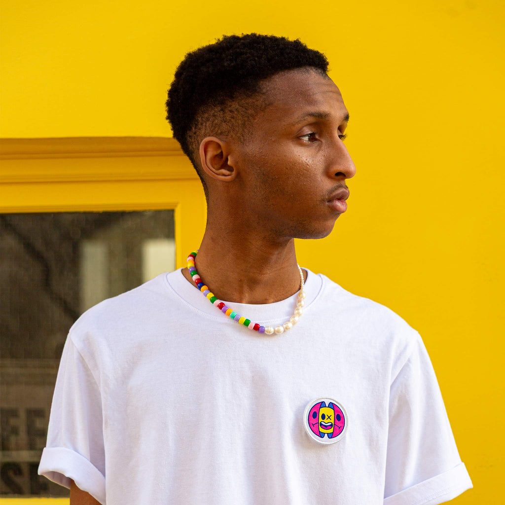 Écusson capsule design porté sur t-shirt blanc par un modèle, devant un mur jaune