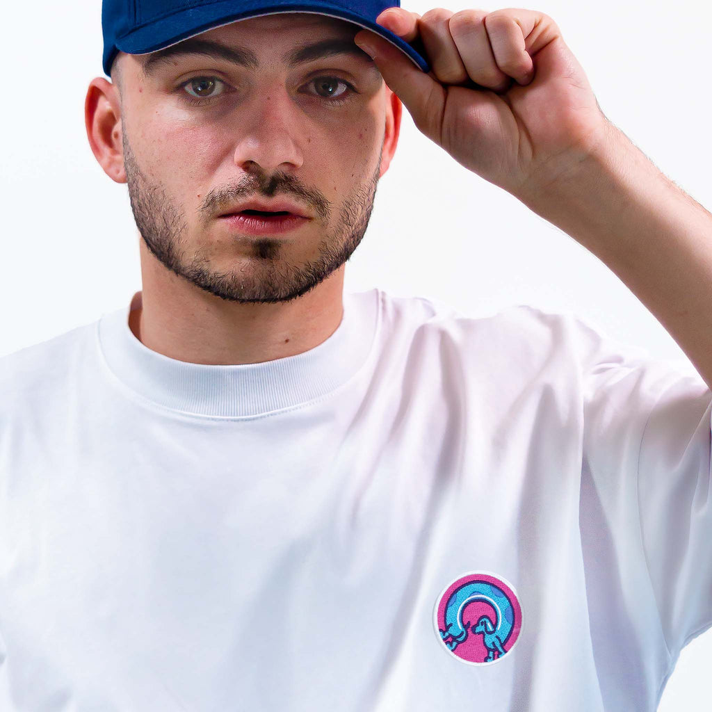 Homme portant un t-shirt blanc avec un patche coloré représentant un chien saucisse, casquette bleue, style moderne.