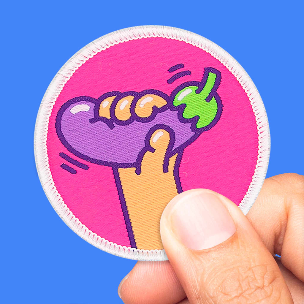 Main tenant un patch rond avec une aubergine violette sur fond rose vif, écusson thermocollant ou scratch.