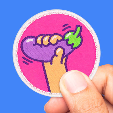 Main tenant un patch rond avec une aubergine violette sur fond rose vif, écusson thermocollant ou scratch.