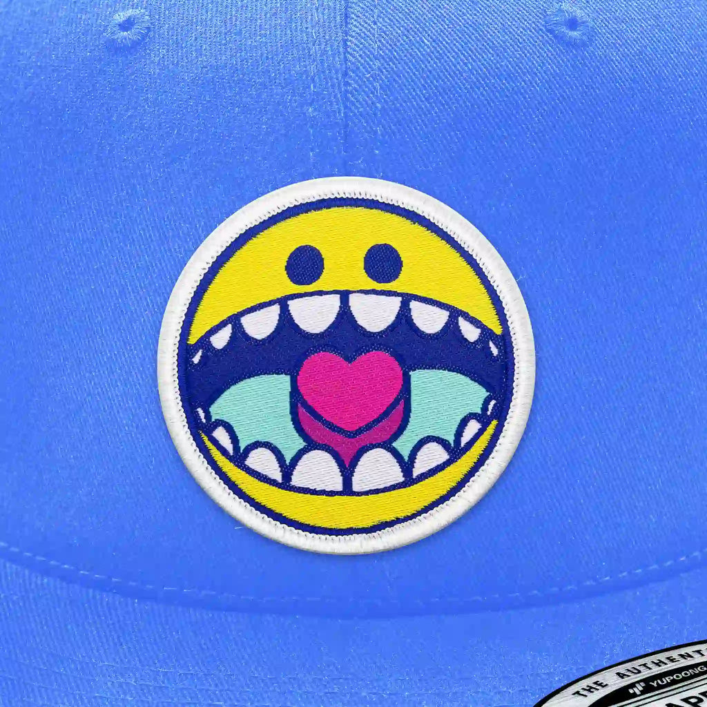 Casquette bleu clair avec écusson thermocollant rond smiley jaune avec grande bouche ouverte et cœur rose, design fun et coloré.