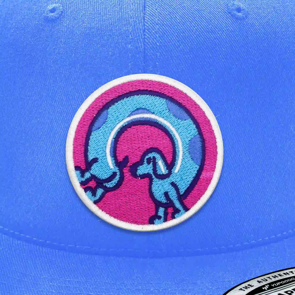 Casquette bleu clair avec écusson thermocollant rond représentant un chien saucisse bleu stylisé sur fond rose et violet.