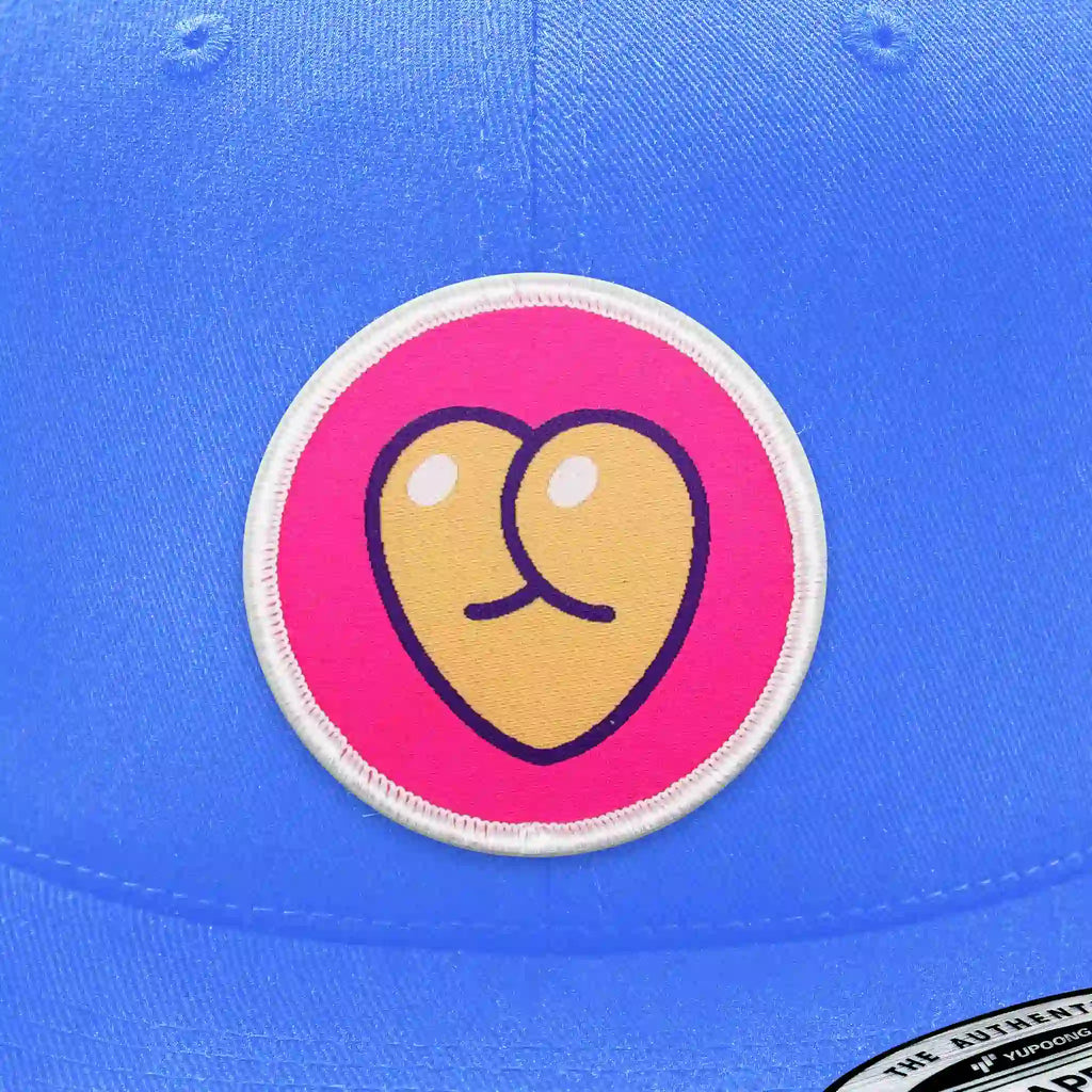 Casquette bleu clair avec écusson thermocollant rond représentant un cœur jaune stylisé en forme de fesses humoristiques.