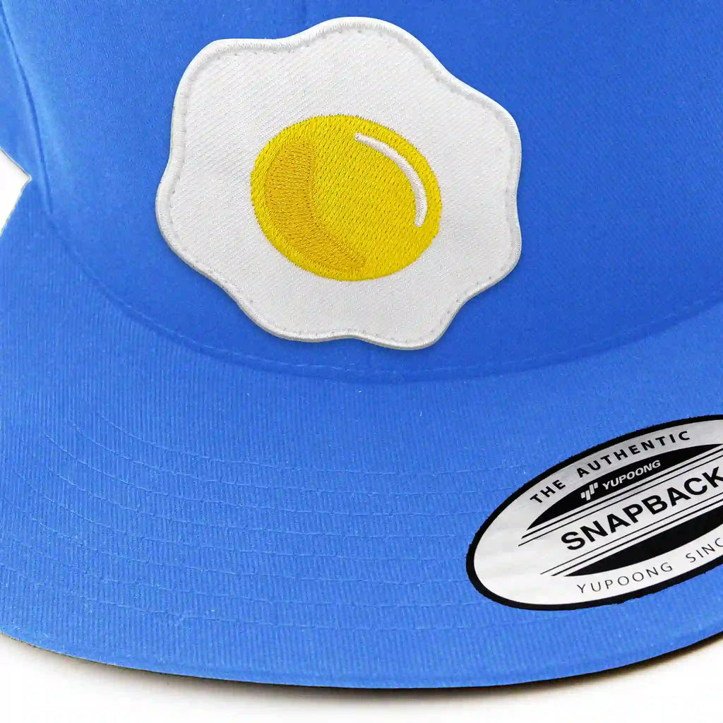 Gros plan sur casquette Flexfit bleu clair avec écusson interchangeable œuf brodé, accessoire streetwear unique.