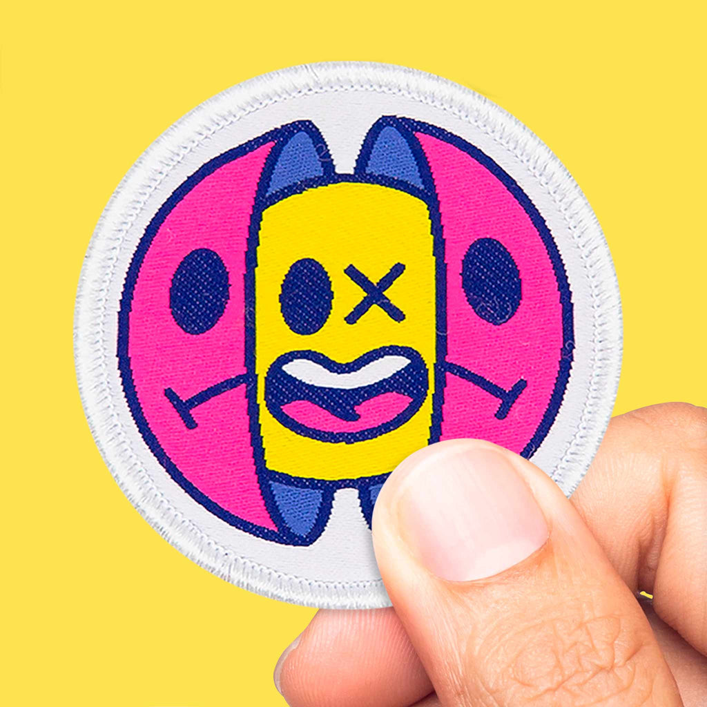 Patch brodé capsule pop art tenu entre les doigts sur fond jaune